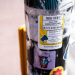 A small advocacy sticker from LUCE Massachusetts is posted to a city pole at Harvard, urging passersby to report ICE sightings and verify enforcement activity through the community hotline. Below it, layers of older stickers and weathered tape add to the textured urban collage—evidence of civic messaging and street culture intersecting. The sticker’s bold yellow and black design stands out against the faded background, capturing a snapshot of immigrant rights activism in a public space.