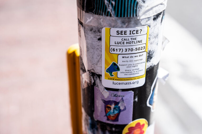 LUCE ICE Watch Sticker, Massachusetts Street Scene 4 A small advocacy sticker from LUCE Massachusetts is posted to a city pole at Harvard, urging passersby to report ICE sightings and verify enforcement activity through the community hotline. Below it, layers of older stickers and weathered tape add to the textured urban collage—evidence of civic messaging and street culture intersecting. The sticker’s bold yellow and black design stands out against the faded background, capturing a snapshot of immigrant rights activism in a public space.