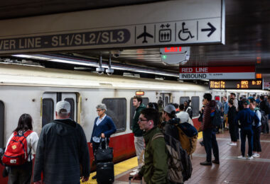 Passengers wait alongside an inbound MBTA Red Line train, headed toward Alewife, on a busy underground platform in downtown Boston. The Red Line is the system’s oldest rapid transit route, opening in 1912 and forming the backbone of east–west and north–south travel through the city. The high-floor cars shown here reflect decades of incremental fleet updates, while the tiled platforms, overhead wayfinding, and digital countdown signs illustrate how the MBTA has layered modern passenger information systems onto early-20th-century station infrastructure. The crowding and luggage visible on the platform highlight the line’s dual role serving both daily commuters and airport-bound travelers via transfers to bus and rail connections.