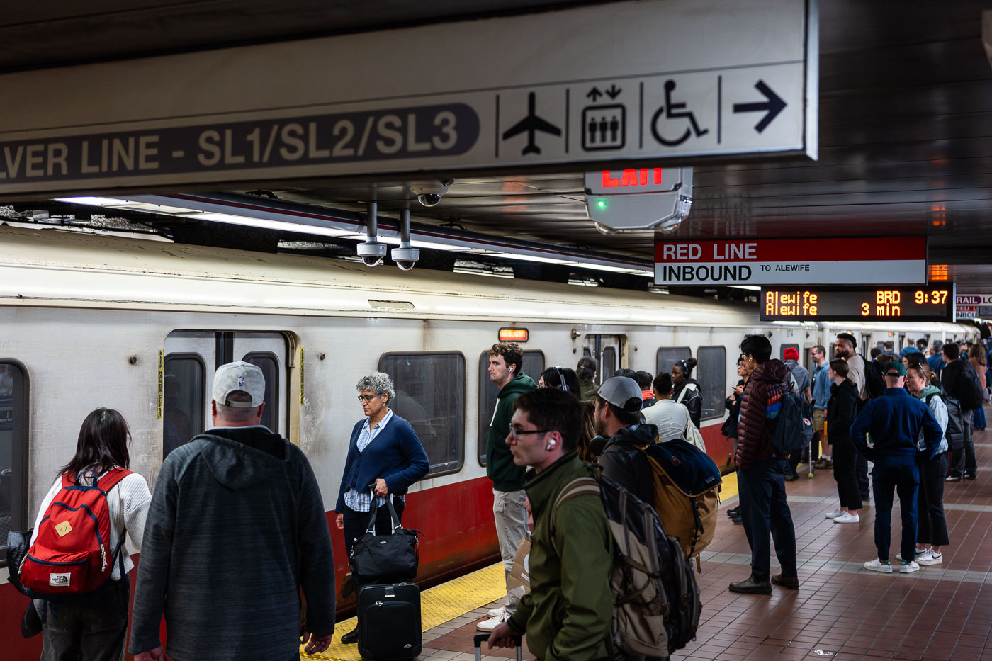 Passengers wait alongside an inbound MBTA Red Line train, headed toward Alewife, on a busy underground platform in downtown Boston. The Red Line is the system’s oldest rapid transit route, opening in 1912 and forming the backbone of east–west and north–south travel through the city. The high-floor cars shown here reflect decades of incremental fleet updates, while the tiled platforms, overhead wayfinding, and digital countdown signs illustrate how the MBTA has layered modern passenger information systems onto early-20th-century station infrastructure. The crowding and luggage visible on the platform highlight the line’s dual role serving both daily commuters and airport-bound travelers via transfers to bus and rail connections.