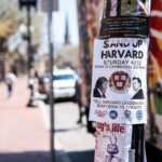 A politically charged flyer near Harvard Square calls for a demonstration at the Cambridge Common, urging the university community to “Stand Up Harvard.” Affixed to a utility pole along Massachusetts Avenue, it features the Harvard crest and contrasting portraits of public figures, symbolizing tension between authority and academic independence. The red-brick sidewalks and blurred foot traffic behind it evoke the constant movement and civic engagement that define this historic Cambridge neighborhood.
