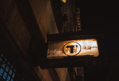 An illuminated MBTA roundel mounted beneath a streetscape, marking an entrance to Boston’s subway system. The simple black “T” inside a white circle has been used in various forms since the mid-20th century and became the unified symbol of the MBTA in the 1960s, replacing a mix of earlier streetcar and rapid-transit branding. Designed for quick recognition in dense urban conditions, the sign’s placement below street level reflects how much of Boston’s transit infrastructure is woven directly into older masonry buildings and narrow rights-of-way. The weathered lightbox and surrounding stonework show decades of exposure, maintenance, and adaptation as the system has evolved while keeping its most recognizable identifier largely unchanged.