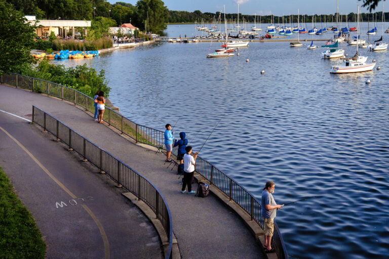 Fishing and Sailboats in Minneapolis 2 People fishing off the shore of Bde Maka Ska, with sailboats on the water.