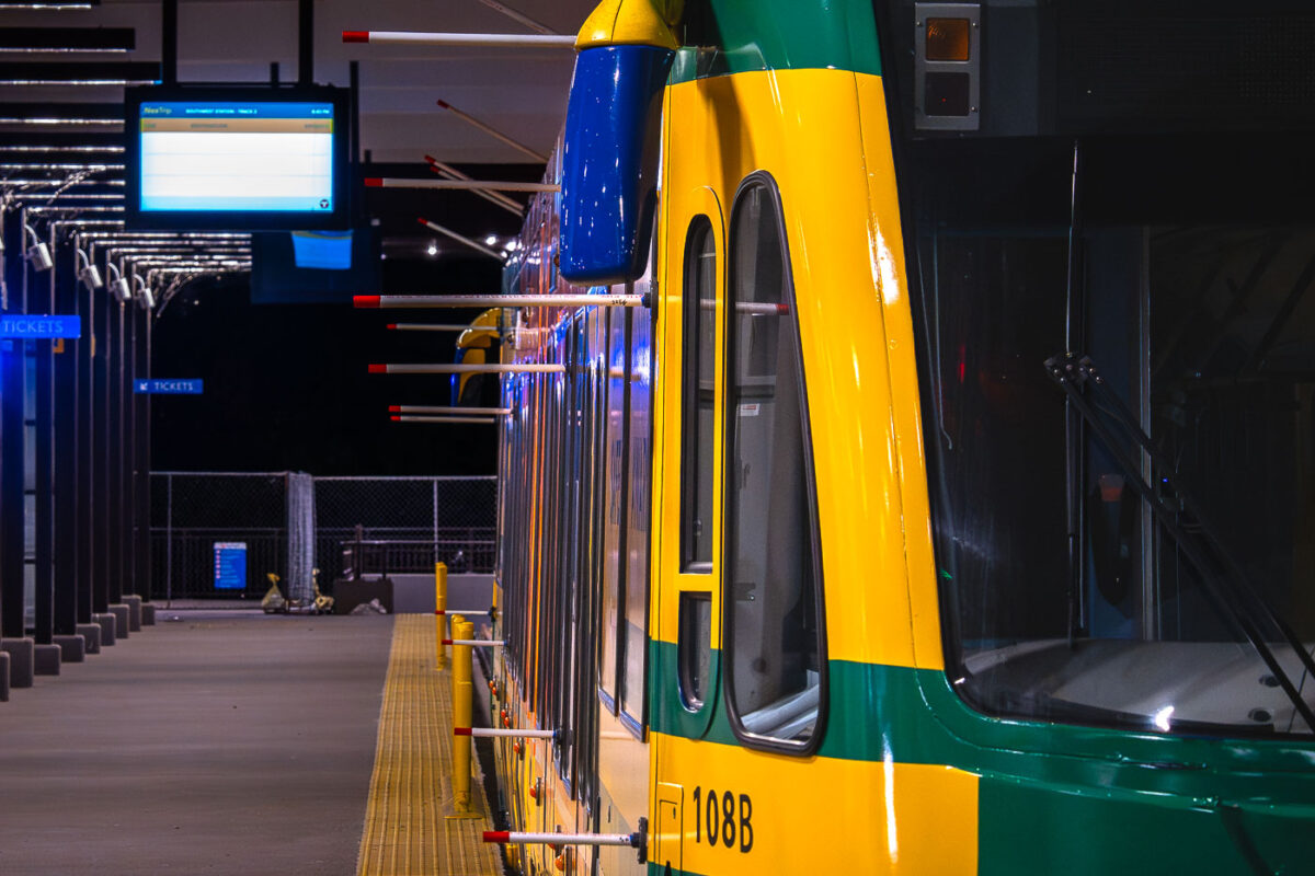 A light rail train, numbered 108B, is stopped at the Southwest Station platform. The station features a digital display indicating "SOUTHWEST STATION: TRACK 2" and "8:50 PM", suggesting it is operational during evening hours. The train's distinctive yellow and green livery is visible, along with safety bollards and ticket signage, indicating a functional public transportation hub. Southwest Station is a key stop on the light rail network, facilitating transit for commuters and visitors within the urban environment.
