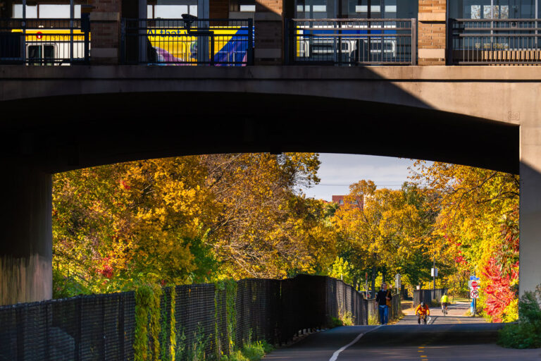Midtown Greenway Colors and Metro Transit above 2 Bikers, walkers on the Midtown Greenway as a bus leaves the Uptown Transit Station above. Uptown Minneapolis, October 2025