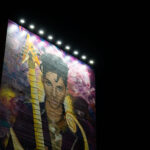 Pricne Mural, Minneapolis 2025 2 A large-scale mural depicting Prince dominates the side of a brick building, illuminated by a row of rooftop lights that emphasize both the portrait and the texture of the masonry beneath it. The artwork presents Prince with his iconic guitar, referencing his role not only as a performer but as a technically accomplished musician and songwriter whose work reshaped popular music beginning in the late 1970s. The saturated purples, golds, and layered background elements visually echo the imagery and symbolism long associated with his career, particularly the Purple Rain era that cemented his global influence.Murals like this became an important part of Minneapolis’s visual landscape following Prince’s death in 2016, serving as public memorials that connect the city’s streets to its musical legacy. Painted directly onto brick, the piece blends fine-art portraiture with urban scale, designed to be read from a distance while still holding detail up close. Night lighting transforms the mural into a landmark after dark, reinforcing Prince’s enduring presence in the city where he was born, recorded, and built an independent creative empire that continues to influence artists across genres.