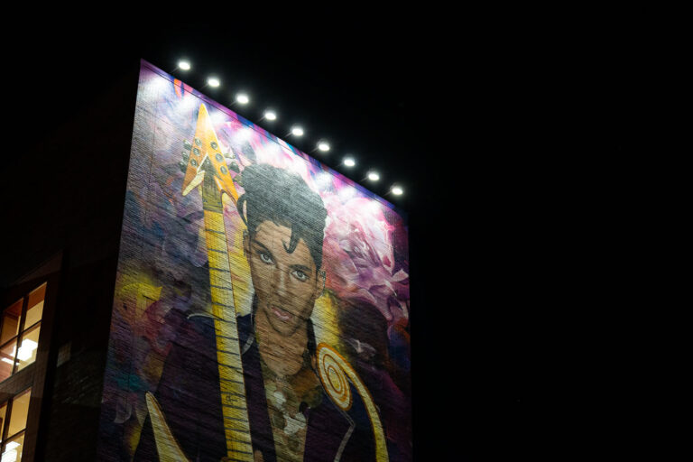 Pricne Mural, Minneapolis 2025 2 A large-scale mural depicting Prince dominates the side of a brick building, illuminated by a row of rooftop lights that emphasize both the portrait and the texture of the masonry beneath it. The artwork presents Prince with his iconic guitar, referencing his role not only as a performer but as a technically accomplished musician and songwriter whose work reshaped popular music beginning in the late 1970s. The saturated purples, golds, and layered background elements visually echo the imagery and symbolism long associated with his career, particularly the Purple Rain era that cemented his global influence.Murals like this became an important part of Minneapolis’s visual landscape following Prince’s death in 2016, serving as public memorials that connect the city’s streets to its musical legacy. Painted directly onto brick, the piece blends fine-art portraiture with urban scale, designed to be read from a distance while still holding detail up close. Night lighting transforms the mural into a landmark after dark, reinforcing Prince’s enduring presence in the city where he was born, recorded, and built an independent creative empire that continues to influence artists across genres.