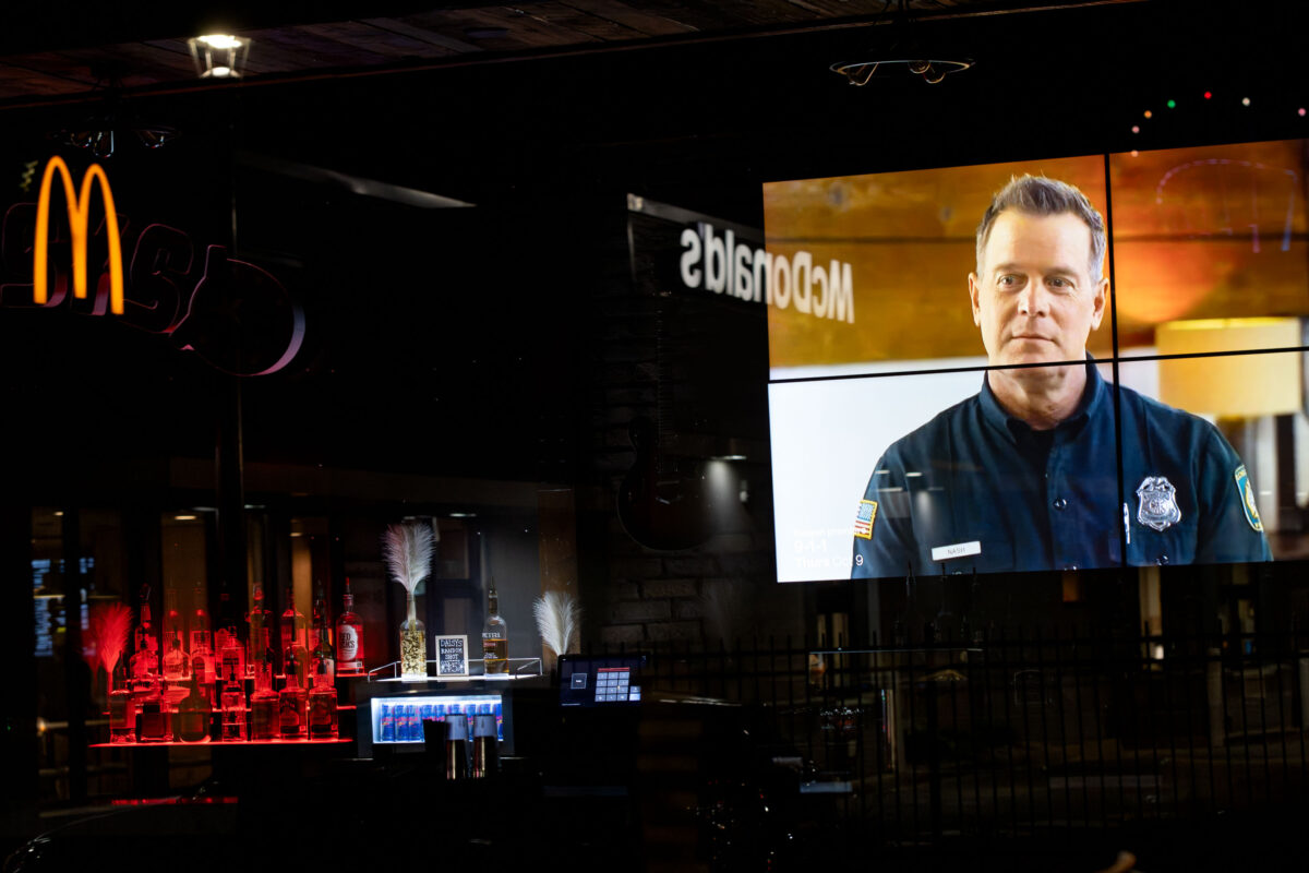 Video ad in a McDonald’s window at night, reflected with the golden arches sign and bar bottles.