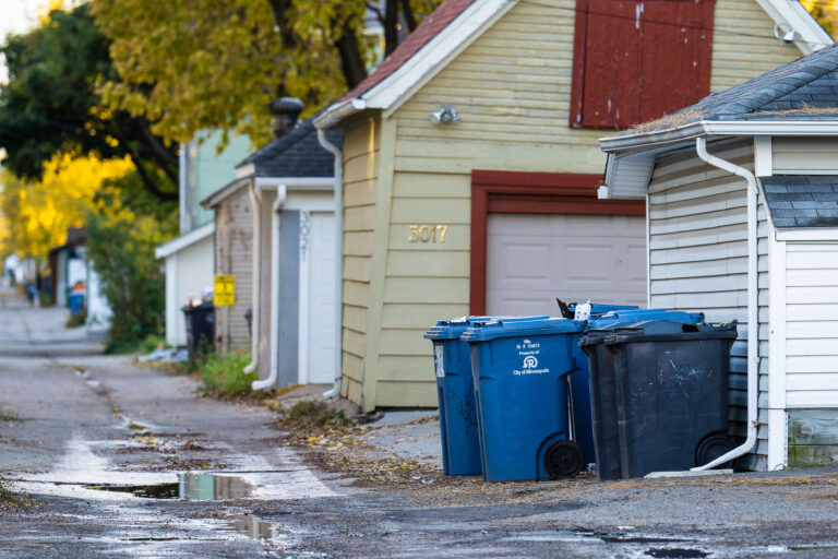 Autumn Alley and Recycling Bins in Uptown Minneapolis 1 A narrow residential alley in Uptown Minneapolis, lined with city-issued recycling and trash bins outside detached garages. The City of Minneapolis operates one of the nation’s oldest organized alley waste collection systems, dating back to the early 20th century when service routes were first formalized for the city’s grid of north-south residential blocks. Today, the alleys continue to serve as vital service corridors, reflecting both the urban infrastructure and the century-old planning model that still defines much of Minneapolis’s residential layout.