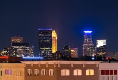 A nighttime view of the Minneapolis skyline as seen from Lake Street in Uptown, looking north toward the city’s central business district. The IDS Center, Minnesota’s tallest building since 1972, anchors the scene on the left with its rectangular black façade and illuminated roofline. Just to the right, the art deco–inspired Wells Fargo Center glows in its signature gold lighting, a design motif rooted in its 1980s postmodern architecture. On the far right, Capella Tower stands out with its distinctive ring-shaped halo, an element added during the 1990s wave of illuminated crown designs across American skyscrapers. The foreground shows the mixed-use buildings of Uptown, reflecting the corridor’s early-20th-century commercial architecture.