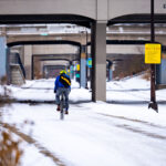 A winter scene along the Midtown Greenway, the 5.5-mile rail-to-trail corridor that runs through the heart of Minneapolis. The trench and bridge structures visible here date back to the early 20th century, when the Milwaukee Road railroad used this grade-separated route to move freight without disrupting street traffic. After rail service declined, the corridor was converted in stages beginning in the late 1990s, becoming one of the city’s most heavily used bicycle and pedestrian routes. The snow-covered path shows how the greenway remains active year-round, with cyclists continuing to use the historic trench even in mid-December.