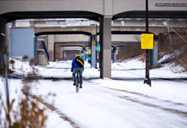 A winter scene along the Midtown Greenway, the 5.5-mile rail-to-trail corridor that runs through the heart of Minneapolis. The trench and bridge structures visible here date back to the early 20th century, when the Milwaukee Road railroad used this grade-separated route to move freight without disrupting street traffic. After rail service declined, the corridor was converted in stages beginning in the late 1990s, becoming one of the city’s most heavily used bicycle and pedestrian routes. The snow-covered path shows how the greenway remains active year-round, with cyclists continuing to use the historic trench even in mid-December.