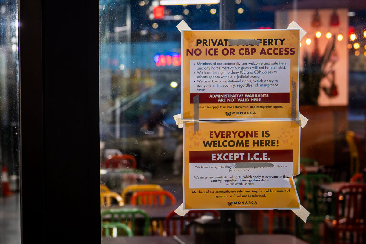 Two printed notices are taped to the front window of a neighborhood business in late December 2025, stating “Private Property — No ICE or CBP Access” and “Everyone Is Welcome Here! Except I.C.E.” The signs outline specific legal language, asserting the right to deny entry to federal immigration authorities without a valid judicial warrant and rejecting the use of administrative warrants.
