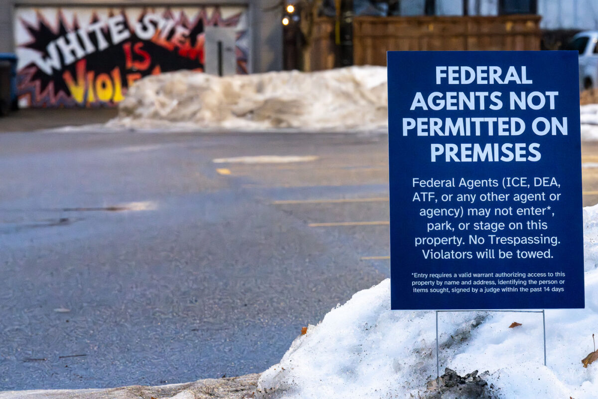 A printed yard sign declaring “Federal Agents Not Permitted on Premises” is planted into a snowbank along a city street in late December 2025. The notice explicitly names agencies such as ICE, the DEA, and ATF, asserting that entry requires a valid, recent warrant and framing the space as protected from routine federal enforcement activity.