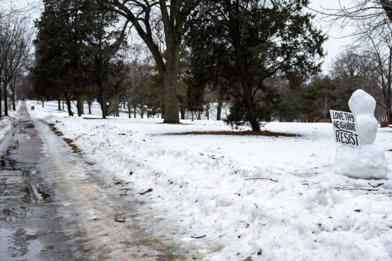 Love Thy Neighbor, Resist — Powderhorn Park 1 A small snow figure stands along a cleared pedestrian path in Powderhorn Park in Minneapolis on December 27, 2025, holding a hand-lettered sign that reads “Love Thy Neighbor — Resist.”