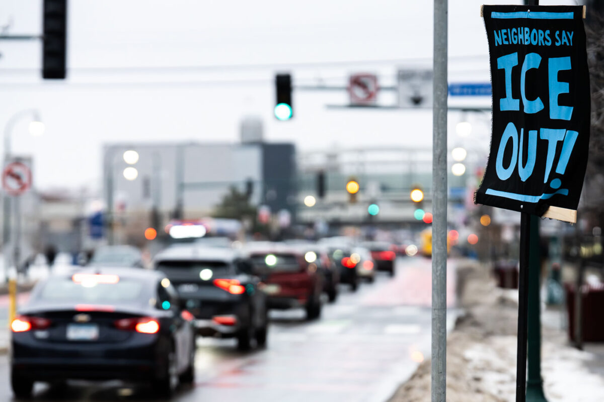 A handmade banner reading “Neighbors Say ICE Out!” is posted along Lake Street in Minneapolis on December 27, 2025, facing steady winter traffic. The message reflects ongoing local opposition to U.S. Immigration and Customs Enforcement, expressed not through an organized demonstration but as a fixed, neighborhood-level statement embedded in daily urban movement.