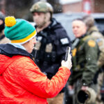 Greg Bovino watches observer blowing whistle 4 An observer blows a whistle at a gas station as Border Patrol commander Gregory Bovino looks on.