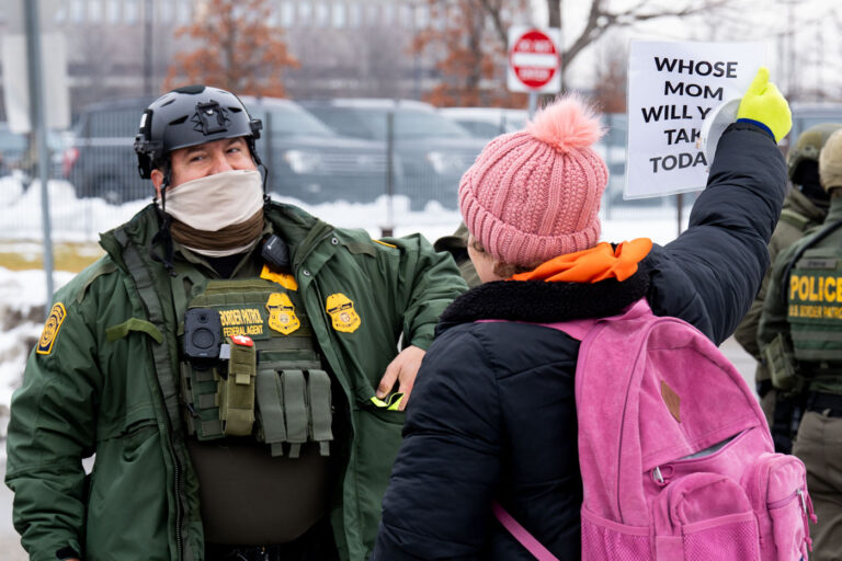 Protester with sign and Border Patrol agent in Minneapolis 2 A protester holds up a sign that reads "Whose mom will you take today?" in front of a Border Patrol Federal Agent.