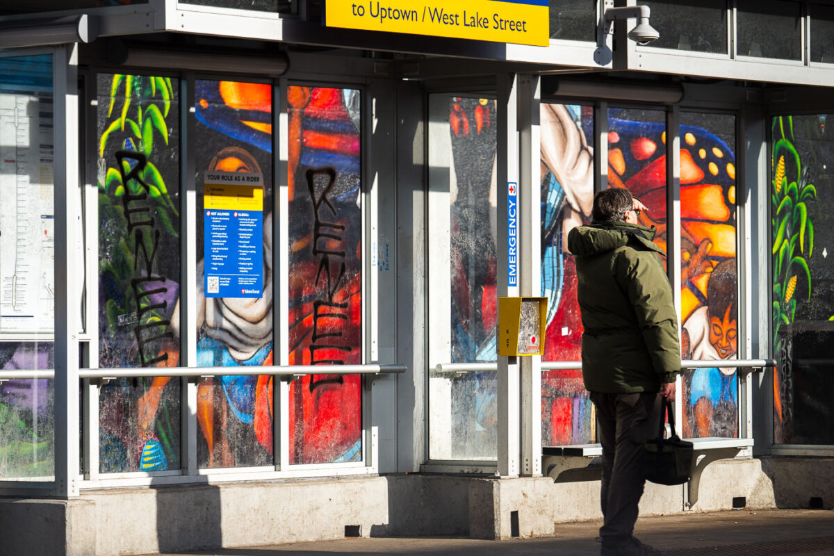 A man stands at a bus shelter with "RENEE" written on the glass windows. Seen at Lake Street in Minneapolis on January 12, 2026.