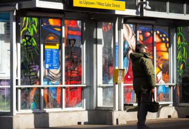 A man stands at a bus shelter with "RENEE" written on the glass windows. Seen at Lake Street in Minneapolis on January 12, 2026.