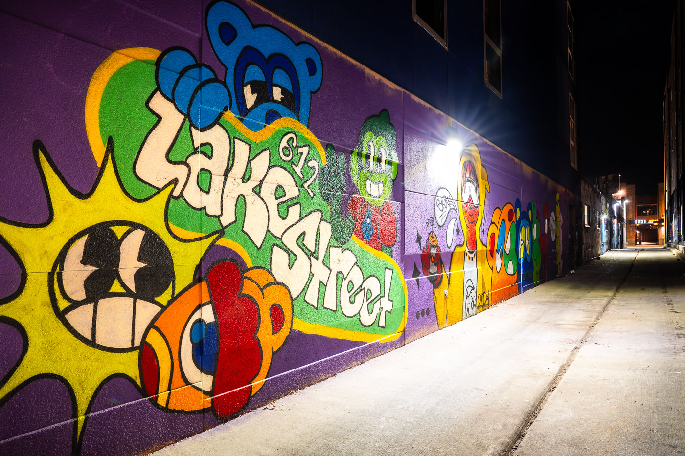 A vibrant mural with cartoon characters and "612 Lake Street" text covers a purple wall in an alleyway in Uptown Minneapolis, MN.