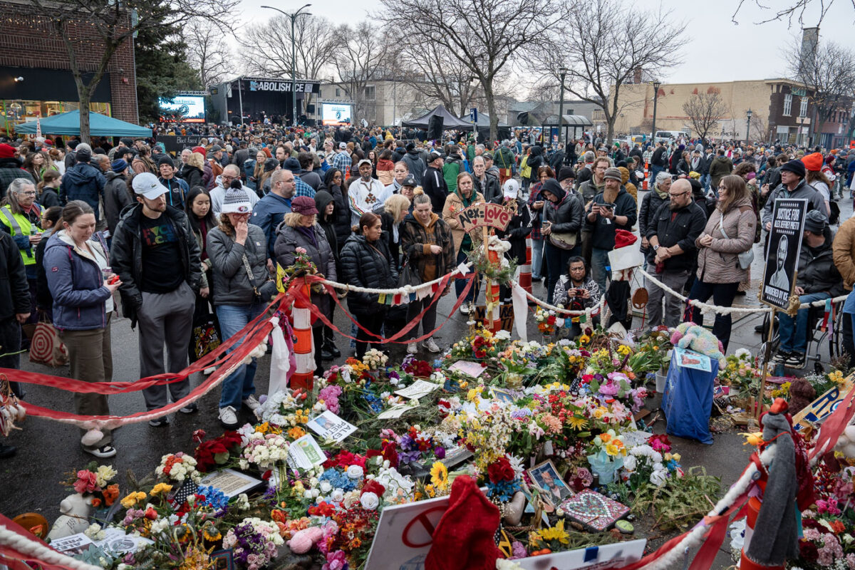A memorial for Alex Pretti, who was "Murdered by ICE," is displayed at a benefit concert for the Abolish ICE movement. The memorial features floral arrangements, photographs, and signs. A stage with "ABOLISH ICE" is visible in the background.
