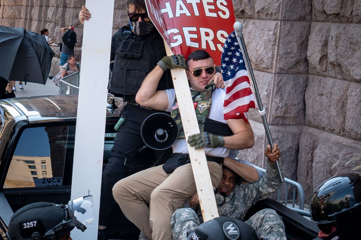 Man in military-style clothing holds megaphone and American flag during confrontation at Minneapolis City Hall.