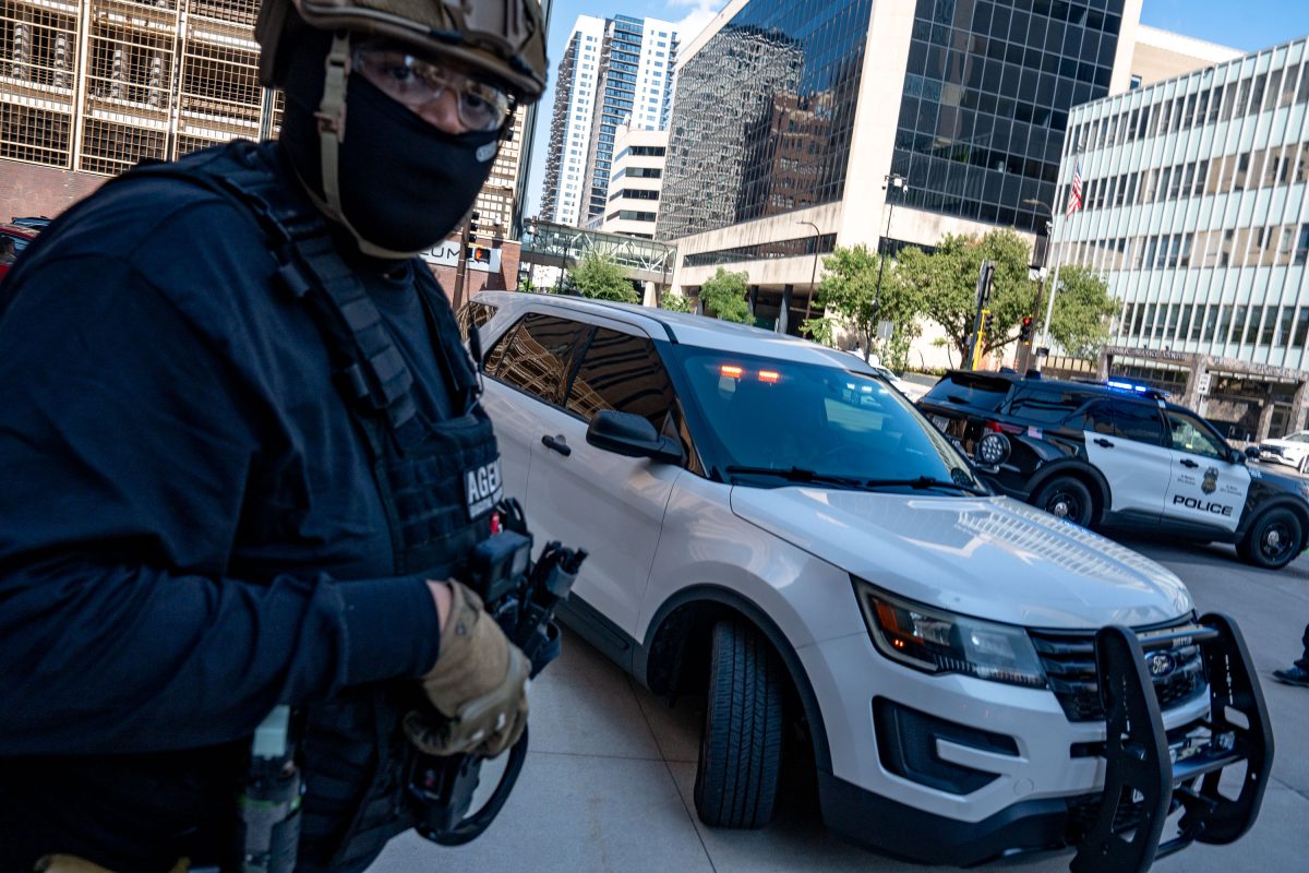 Police officer in tactical gear stands near white police vehicle during downtown Minneapolis protest.