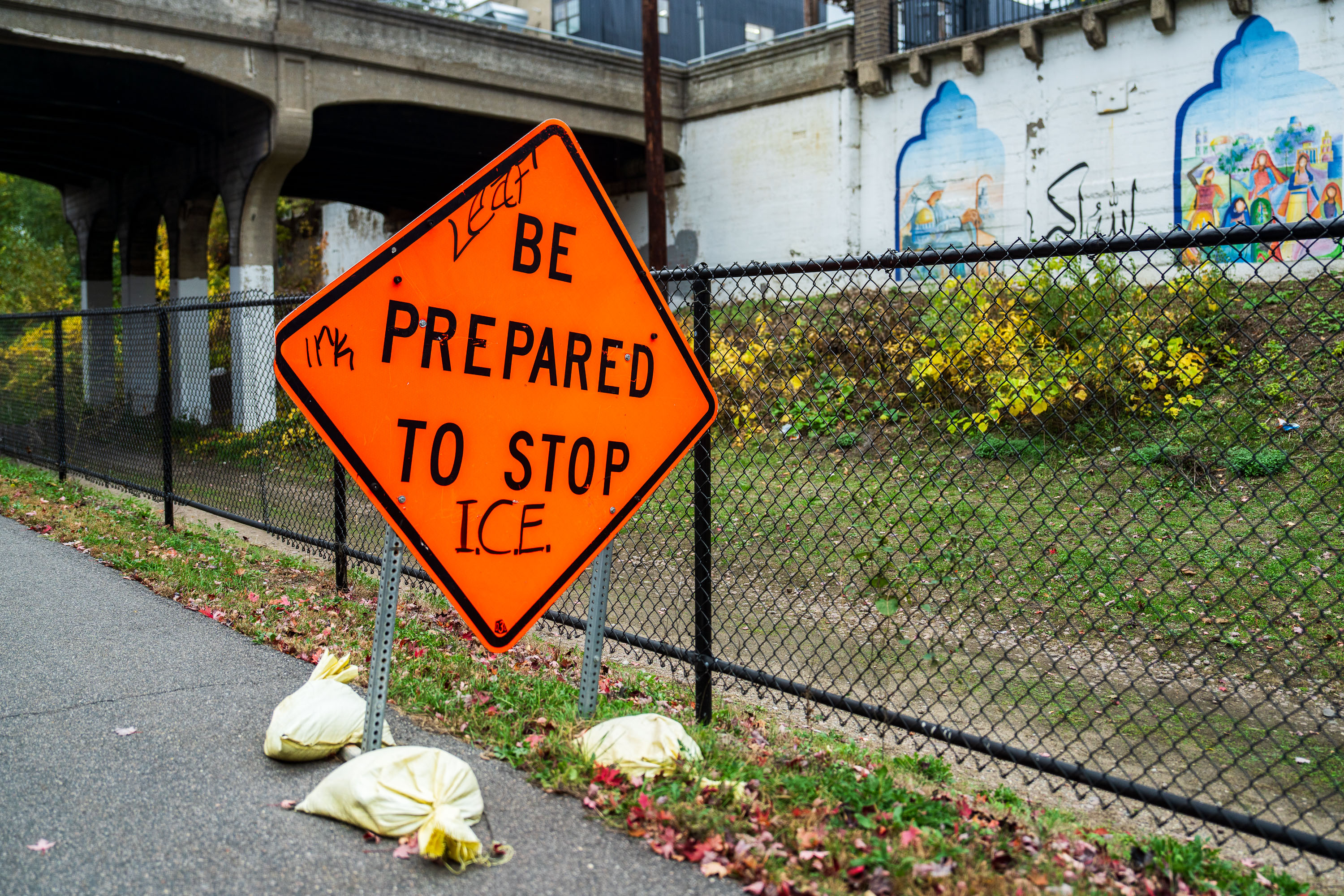 Orange diamond-shaped sign reading "BE PREPARED TO STOP ICE" mounted on fence along Midtown Greenway.