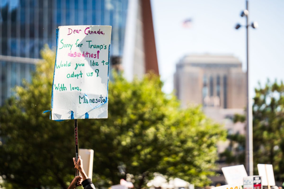 Protester holds handmade sign addressed to Canada apologizing for Trump's policies and asking for adoption at the No Kings rally in Minneapolis Commons Park.