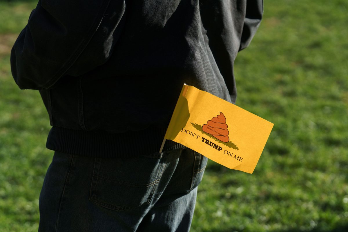 Person holding a yellow flag with a poop emoji and "Don't Trump On Me" text at an outdoor protest.