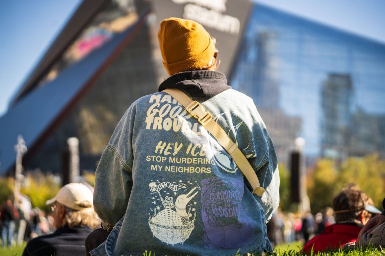 "Hey ICE, Stop Murdering My Neighbors" Jacket at the No Kings Rally 2 A protester wearing a yellow beanie and messaging jacket stands at the NO KINGS rally in Commons Park, Minneapolis.