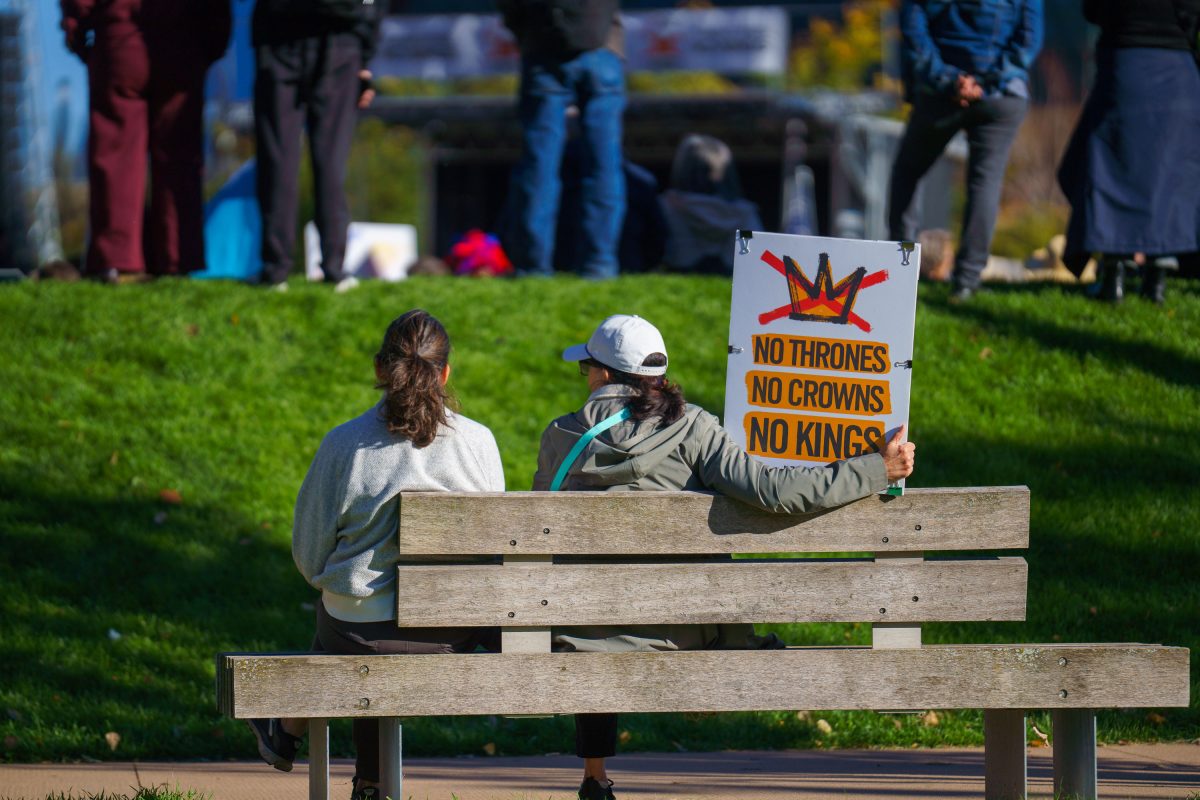 Two people sit on a bench holding a protest sign reading "No Thrones No Crowns No Kings" at a Minneapolis rally.