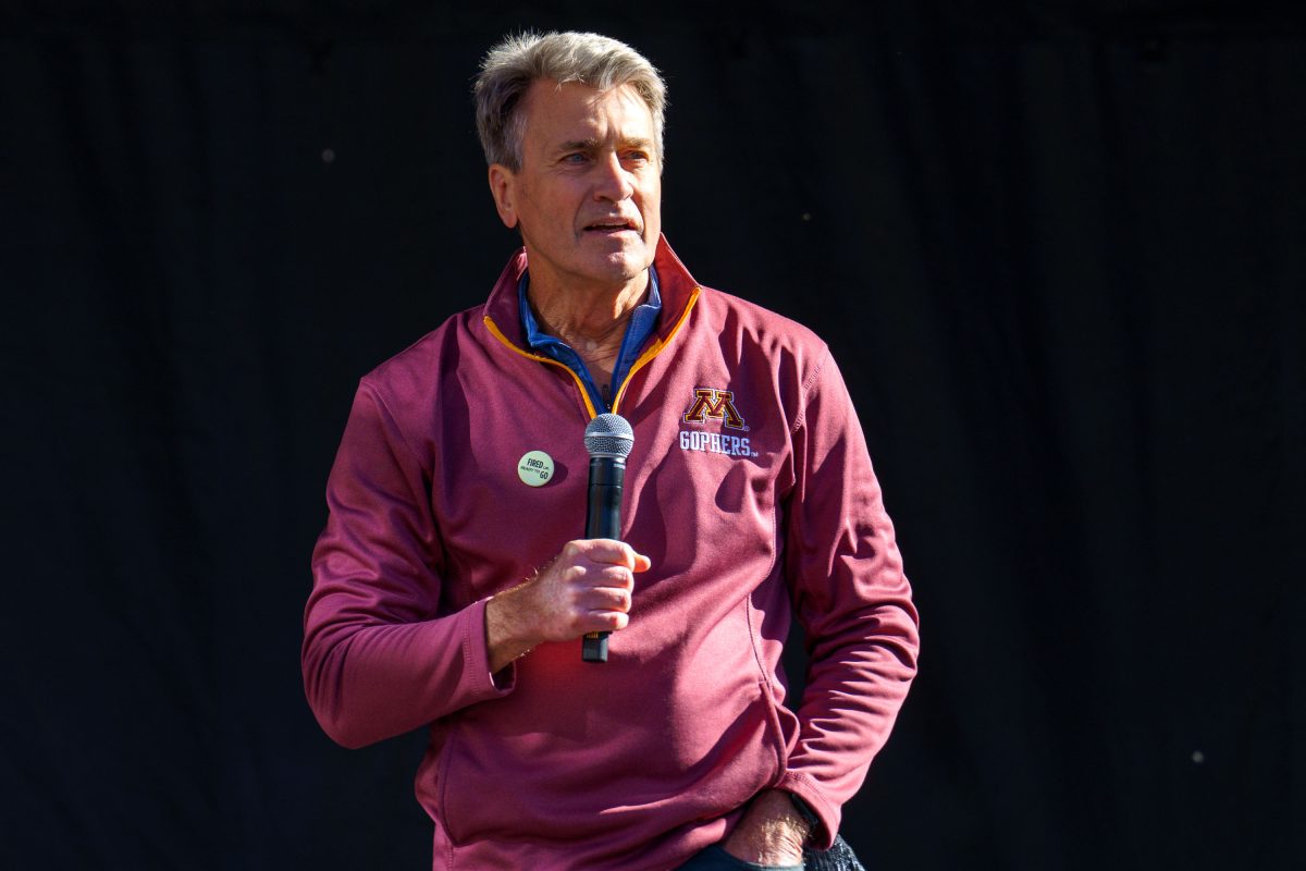 A man in a maroon jacket holding a microphone speaks at a Minneapolis rally or protest event.
