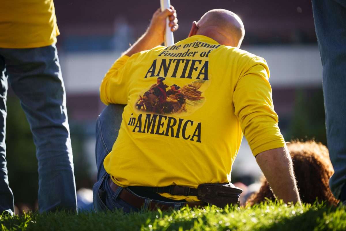 Person wearing yellow shirt reading "The original Founder of ANTIFA in AMERICA" kneels on grass during protest.