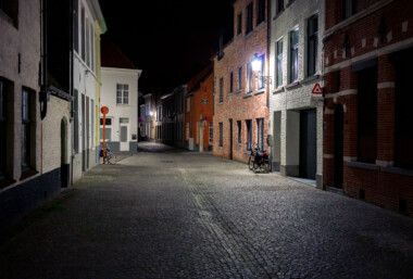 A quiet cobblestone lane in the historic center of Bruges, Belgium, stands nearly empty at night. Rows of brick and plaster townhouses line the narrow street, their windows dark, while a wall-mounted streetlamp casts light on a pair of parked bicycles. Standard European road signs are visible, including a red no‑entry sign at left and a triangular warning sign indicating cycle traffic on the right. Bruges’ medieval core, a UNESCO World Heritage site, retains many car‑restricted streets where bicycles are a common mode of transport for residents and visitors. The scene reflects the city’s emphasis on preserving historic architecture and prioritizing slow, local traffic in its dense urban fabric. A quiet cobblestone lane in the historic center of Bruges, Belgium, stands nearly empty at night. Rows of brick and plaster townhouses line the narrow street, their windows dark, while a wall-mounted streetlamp casts light on a pair of parked bicycles. Standard European road signs are visible, including a red no‑entry sign at left and a triangular warning sign indicating cycle traffic on the right. Bruges’ medieval core, a UNESCO World Heritage site, retains many car‑restricted streets where bicycles are a common mode of transport for residents and visitors. The scene reflects the city’s emphasis on preserving historic architecture and prioritizing slow, local traffic in its dense urban fabric.