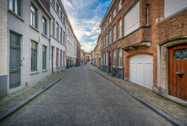 A quiet residential lane in Bruges, Belgium, stretches between rows of brick and plastered townhouses. Cobblestones pave the narrow street, which is lined with ground‑floor doors, shuttered windows, and a white arched garage entrance on the right. Several bicycles are parked against the façades, and a small traffic sign is visible toward the intersection in the distance. The scene sits within Bruges’ historic center, a UNESCO World Heritage site known for its well‑preserved medieval street grid and domestic architecture. Daytime clouds drift over the low‑rise buildings, and no pedestrians or vehicles are present. A quiet residential lane in Bruges, Belgium, stretches between rows of brick and plastered townhouses. Cobblestones pave the narrow street, which is lined with ground‑floor doors, shuttered windows, and a white arched garage entrance on the right. Several bicycles are parked against the façades, and a small traffic sign is visible toward the intersection in the distance. The scene sits within Bruges’ historic center, a UNESCO World Heritage site known for its well‑preserved medieval street grid and domestic architecture. Daytime clouds drift over the low‑rise buildings, and no pedestrians or vehicles are present.