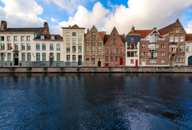 Stepped-gable townhouses line a canal of the River Reie in central Bruges, Belgium. A row of brick and plaster façades, some medieval in style and others with modern additions, faces the water across a stone quay. A Belgian tricolor flag hangs from a doorway at left, while red-painted doors and window frames punctuate several of the buildings. Bruges’ historic center, a UNESCO World Heritage Site since 2000, preserves the canal network that once linked this former medieval trading hub to the North Sea. No pedestrians or boats are visible on the quiet waterfront under partly cloudy skies. Keywords — Activities: sightseeing, canal-side walking; Buildings: stepped-gable townhouses, guildhouses, apartments; Location: Bruges, West Flanders, Belgium, River Reie; Objects: Belgian flag, windows, doors, balconies, stone quay; People: none visible; Moods: tranquil; Sceneries: urban waterfront; Texts: traffic signs; Companies: none; Weather: partly cloudy; Plants: none; Animals: none; Vehicles: none. Stepped-gable townhouses line a canal of the River Reie in central Bruges, Belgium. A row of brick and plaster façades, some medieval in style and others with modern additions, faces the water across a stone quay. A Belgian tricolor flag hangs from a doorway at left, while red-painted doors and window frames punctuate several of the buildings. Bruges’ historic center, a UNESCO World Heritage Site since 2000, preserves the canal network that once linked this former medieval trading hub to the North Sea. No pedestrians or boats are visible on the quiet waterfront under partly cloudy skies. Keywords — Activities: sightseeing, canal-side walking; Buildings: stepped-gable townhouses, guildhouses, apartments; Location: Bruges, West Flanders, Belgium, River Reie; Objects: Belgian flag, windows, doors, balconies, stone quay; People: none visible; Moods: tranquil; Sceneries: urban waterfront; Texts: traffic signs; Companies: none; Weather: partly cloudy; Plants: none; Animals: none; Vehicles: none.