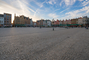 Evening on the Markt, Bruges, Belgium. Visitors cross the wide cobblestoned square as sunset light outlines the row of step‑gabled guild houses that front cafés with green awnings. At the center stands the 1887 monument to Jan Breydel and Pieter de Coninck, ringed by flagpoles and a cast‑iron lamppost. The scene captures a typical early evening in Bruges’ historic core, a UNESCO‑listed medieval city where the Markt has hosted trade and civic life for centuries. Contrails and thin clouds streak the sky while small groups linger near bicycles and restaurant terraces. Keywords- Activities: strolling, sightseeing, cycling, dining- Buildings: step-gabled guild houses, cafés, monument plinths- Location: Bruges, West Flanders, Belgium; Market Square (Markt)- Objects: cobblestones, lamppost, flagpoles, café awnings, clock- People: tourists, locals, pedestrians- Moods: calm, unhurried, early evening- Sceneries: historic town square, open plaza- Texts: storefront signage, building numbers (unreadable at distance)- Companies: local cafés and restaurants (unidentified)- Weather: clear with high clouds at sunset- Plants: none visible- Animals: none visible- Vehicles: bicycles Evening on the Markt, Bruges, Belgium. Visitors cross the wide cobblestoned square as sunset light outlines the row of step‑gabled guild houses that front cafés with green awnings. At the center stands the 1887 monument to Jan Breydel and Pieter de Coninck, ringed by flagpoles and a cast‑iron lamppost. The scene captures a typical early evening in Bruges’ historic core, a UNESCO‑listed medieval city where the Markt has hosted trade and civic life for centuries. Contrails and thin clouds streak the sky while small groups linger near bicycles and restaurant terraces. Keywords- Activities: strolling, sightseeing, cycling, dining- Buildings: step-gabled guild houses, cafés, monument plinths- Location: Bruges, West Flanders, Belgium; Market Square (Markt)- Objects: cobblestones, lamppost, flagpoles, café awnings, clock- People: tourists, locals, pedestrians- Moods: calm, unhurried, early evening- Sceneries: historic town square, open plaza- Texts: storefront signage, building numbers (unreadable at distance)- Companies: local cafés and restaurants (unidentified)- Weather: clear with high clouds at sunset- Plants: none visible- Animals: none visible- Vehicles: bicycles