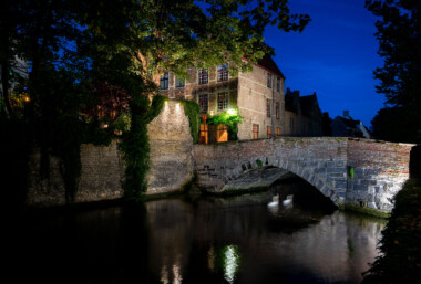 Medieval bridge on the Reie, Bruges at dusk. A single-arch stone and brick bridge spans a quiet bend of the River Reie in Bruges, Belgium, its parapet and adjoining quay lit by warm lamps that reflect on the canal. Behind the bridge, ivy climbs weathered brick facades and gabled townhouses typical of the city’s historic core, a UNESCO World Heritage site. Leafy trees frame the scene, and the waterway shows faint ripples but no pedestrians or boats are visible. The location sits along one of Bruges’ centuries-old canals that once supported trade and still anchor the city’s tourism and daily life. Keywords — Activities: evening strolls, sightseeing; Buildings: historic townhouses, canal quay; Location: Bruges, Belgium, River Reie, historic center; Objects: streetlights, ivy-covered walls, windows; People: none visible; Moods: tranquil, contemplative; Sceneries: canal, old bridge, reflections; Texts: none; Companies: none; Weather: clear evening; Plants: trees, ivy; Animals: none visible; Vehicles: none visible. Medieval bridge on the Reie, Bruges at dusk. A single-arch stone and brick bridge spans a quiet bend of the River Reie in Bruges, Belgium, its parapet and adjoining quay lit by warm lamps that reflect on the canal. Behind the bridge, ivy climbs weathered brick facades and gabled townhouses typical of the city’s historic core, a UNESCO World Heritage site. Leafy trees frame the scene, and the waterway shows faint ripples but no pedestrians or boats are visible. The location sits along one of Bruges’ centuries-old canals that once supported trade and still anchor the city’s tourism and daily life. Keywords — Activities: evening strolls, sightseeing; Buildings: historic townhouses, canal quay; Location: Bruges, Belgium, River Reie, historic center; Objects: streetlights, ivy-covered walls, windows; People: none visible; Moods: tranquil, contemplative; Sceneries: canal, old bridge, reflections; Texts: none; Companies: none; Weather: clear evening; Plants: trees, ivy; Animals: none visible; Vehicles: none visible.