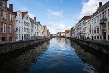 River Reie canal through Bruges’ historic center. Rows of brick and whitewashed townhouses with stepped gables line both banks of the River Reie in Bruges, Belgium. A Belgian tricolor flag hangs from one façade, and wrought-iron street lamps and balconies frame tall windows along the quay. Several pedestrians walk the right-hand embankment as calm water reflects the buildings and a low arched bridge visible in the distance. The scene is photographed in daylight under a partly cloudy sky within the UNESCO-listed medieval core known for its preserved canal network. Keywords — Activities: walking, sightseeing; Buildings: gabled townhouses, historic façades; Location: Bruges, Belgium, River Reie, historic center; Objects: Belgian flag, street lamps, balconies, canal walls; People: pedestrians, tourists; Moods: calm, reflective; Sceneries: canal, waterfront, bridge; Texts: none visible; Companies: none; Weather: partly cloudy, fair; Plants: sparse greenery on quay; Animals: none; Vehicles: none. River Reie canal through Bruges’ historic center. Rows of brick and whitewashed townhouses with stepped gables line both banks of the River Reie in Bruges, Belgium. A Belgian tricolor flag hangs from one façade, and wrought-iron street lamps and balconies frame tall windows along the quay. Several pedestrians walk the right-hand embankment as calm water reflects the buildings and a low arched bridge visible in the distance. The scene is photographed in daylight under a partly cloudy sky within the UNESCO-listed medieval core known for its preserved canal network. Keywords — Activities: walking, sightseeing; Buildings: gabled townhouses, historic façades; Location: Bruges, Belgium, River Reie, historic center; Objects: Belgian flag, street lamps, balconies, canal walls; People: pedestrians, tourists; Moods: calm, reflective; Sceneries: canal, waterfront, bridge; Texts: none visible; Companies: none; Weather: partly cloudy, fair; Plants: sparse greenery on quay; Animals: none; Vehicles: none.