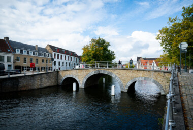 Pedestrians cross a three-arched brick bridge over a branch of the River Reie in Bruges, Belgium. Historic row houses with red-tiled roofs and a mix of gabled and flat façades line the canal, while bollards and railings mark the waterway and walkway. Trees showing early autumn color frame the scene, and a partly cloudy sky brightens the medieval streetscape. The canal network, once the commercial lifeline of the city, is part of the UNESCO-listed Historic Centre of Brugge and remains a focal point for daily life and tourism. Pedestrians cross a three-arched brick bridge over a branch of the River Reie in Bruges, Belgium. Historic row houses with red-tiled roofs and a mix of gabled and flat façades line the canal, while bollards and railings mark the waterway and walkway. Trees showing early autumn color frame the scene, and a partly cloudy sky brightens the medieval streetscape. The canal network, once the commercial lifeline of the city, is part of the UNESCO-listed Historic Centre of Brugge and remains a focal point for daily life and tourism.