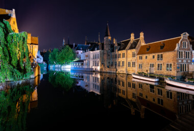 Nighttime reflections line a bend of the River Reie in Bruges, Belgium, as historic brick guild houses and stepped-gable facades mirror perfectly on the still canal. Two white tour boats sit moored for the night along the quay, while a vine-draped wall and a waterside restaurant pavilion are illuminated across the water. Church spires rise in silhouette behind the row of houses, anchoring the skyline of the medieval core. Bruges’ historic center, a UNESCO World Heritage Site, is known for its preserved canals and architecture that draw heavy daytime tourism; after hours, the waterways fall quiet except for lights from hotels and eateries along the quay. Nighttime reflections line a bend of the River Reie in Bruges, Belgium, as historic brick guild houses and stepped-gable facades mirror perfectly on the still canal. Two white tour boats sit moored for the night along the quay, while a vine-draped wall and a waterside restaurant pavilion are illuminated across the water. Church spires rise in silhouette behind the row of houses, anchoring the skyline of the medieval core. Bruges’ historic center, a UNESCO World Heritage Site, is known for its preserved canals and architecture that draw heavy daytime tourism; after hours, the waterways fall quiet except for lights from hotels and eateries along the quay.