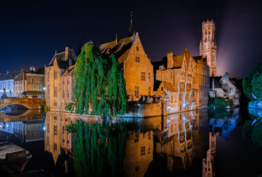 Bruges’ medieval waterfront is illuminated at night along the River Reie in the historic center of Bruges, Belgium. Brick gabled houses line the canal, their facades reflected in still water beside a stone arch bridge at left and moored boats in the foreground. A cascading willow tree drapes over the quay, while cafe terraces sit behind a low brick wall. In the background, the Belfry of Bruges rises above the rooftops, its 13th‑century tower lit from behind. The UNESCO‑listed old town’s canals, once part of a medieval trading network, are a focal point for visitors and local commerce throughout the year. Bruges’ medieval waterfront is illuminated at night along the River Reie in the historic center of Bruges, Belgium. Brick gabled houses line the canal, their facades reflected in still water beside a stone arch bridge at left and moored boats in the foreground. A cascading willow tree drapes over the quay, while cafe terraces sit behind a low brick wall. In the background, the Belfry of Bruges rises above the rooftops, its 13th‑century tower lit from behind. The UNESCO‑listed old town’s canals, once part of a medieval trading network, are a focal point for visitors and local commerce throughout the year.