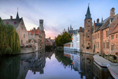 Rozenhoedkaai at dusk, Bruges, Belgium. Historic brick guildhouses and stepped gables line the River Reie, their facades and spires reflected in the still canal water. The Belfry of Bruges rises in the background, while a round turret and narrow townhouses frame the right bank. Covered canal tour boats are moored in the foreground, and a café terrace on the left hosts diners under umbrellas. This vantage, among the city’s most photographed, sits within the UNESCO-listed medieval core of Bruges in West Flanders, a reminder of the trading wealth that shaped the canal network.Keywords — Activities: dining, sightseeing, canal tours; Buildings: medieval guildhouses, Belfry of Bruges, turreted townhouses; Location: Bruges, West Flanders, Belgium, Rozenhoedkaai, River Reie; Objects: umbrellas, café tables, red sign, mooring posts; People: diners, pedestrians; Moods: tranquil, reflective; Sceneries: canal, historic cityscape; Texts: signage on canal-side building; Companies: none visible; Weather: clear, calm evening; Plants: weeping willow, canal-side trees; Animals: none visible; Vehicles: tour boats. Rozenhoedkaai at dusk, Bruges, Belgium. Historic brick guildhouses and stepped gables line the River Reie, their facades and spires reflected in the still canal water. The Belfry of Bruges rises in the background, while a round turret and narrow townhouses frame the right bank. Covered canal tour boats are moored in the foreground, and a café terrace on the left hosts diners under umbrellas. This vantage, among the city’s most photographed, sits within the UNESCO-listed medieval core of Bruges in West Flanders, a reminder of the trading wealth that shaped the canal network.Keywords — Activities: dining, sightseeing, canal tours; Buildings: medieval guildhouses, Belfry of Bruges, turreted townhouses; Location: Bruges, West Flanders, Belgium, Rozenhoedkaai, River Reie; Objects: umbrellas, café tables, red sign, mooring posts; People: diners, pedestrians; Moods: tranquil, reflective; Sceneries: canal, historic cityscape; Texts: signage on canal-side building; Companies: none visible; Weather: clear, calm evening; Plants: weeping willow, canal-side trees; Animals: none visible; Vehicles: tour boats.