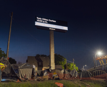 Billboard stating "Twin Cities Curfew. Please stay home" with a homeless camp below. Shot during the 2020 Minneapolis unrest over the May 25th death of George Floyd. Billboard stating "Twin Cities Curfew. Please stay home" with a homeless camp below. Shot during the 2020 Minneapolis unrest over the May 25th death of George Floyd.