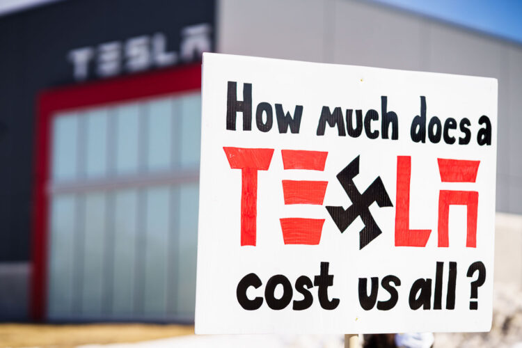 Protester holds a sign that reads "How much does Tesla cost us all?". Seen at a Tesla facility just outside of Minneapolis (Golden Valley) on March 8, 2025.This is the 4th week in a row protesters have gathered here as part of the nationwide Tesla Takedown organizing. Protester holds a sign that reads "How much does Tesla cost us all?". Seen at a Tesla facility just outside of Minneapolis (Golden Valley) on March 8, 2025.This is the 4th week in a row protesters have gathered here as part of the nationwide Tesla Takedown organizing.