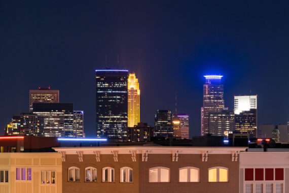 A nighttime view of the Minneapolis skyline as seen from Lake Street in Uptown, looking north toward the city’s central business district. The IDS Center, Minnesota’s tallest building since 1972, anchors the scene on the left with its rectangular black façade and illuminated roofline. Just to the right, the art deco–inspired Wells Fargo Center glows in its signature gold lighting, a design motif rooted in its 1980s postmodern architecture. On the far right, Capella Tower stands out with its distinctive ring-shaped halo, an element added during the 1990s wave of illuminated crown designs across American skyscrapers. The foreground shows the mixed-use buildings of Uptown, reflecting the corridor’s early-20th-century commercial architecture. A nighttime view of the Minneapolis skyline as seen from Lake Street in Uptown, looking north toward the city’s central business district. The IDS Center, Minnesota’s tallest building since 1972, anchors the scene on the left with its rectangular black façade and illuminated roofline. Just to the right, the art deco–inspired Wells Fargo Center glows in its signature gold lighting, a design motif rooted in its 1980s postmodern architecture. On the far right, Capella Tower stands out with its distinctive ring-shaped halo, an element added during the 1990s wave of illuminated crown designs across American skyscrapers. The foreground shows the mixed-use buildings of Uptown, reflecting the corridor’s early-20th-century commercial architecture.